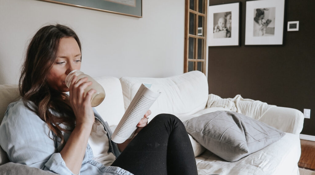 Christian homemaker sits on white couch drinking a coffee and reading a book